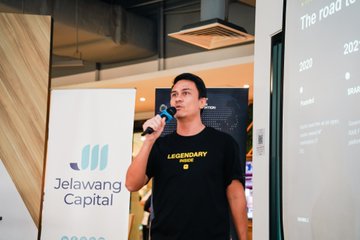 A man wearing a black 'LEGENDARY' t-shirt speaks into a microphone at an event, standing beside a 'Jelawang Capital' banner.