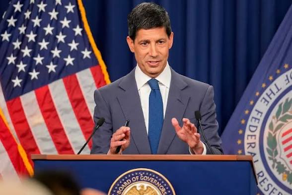 A man in a gray suit and blue tie speaks at a podium bearing the 'BOARD OF GOVERNORS OF THE FEDERAL RESERVE SYSTEM' seal, with the American flag behind him.
