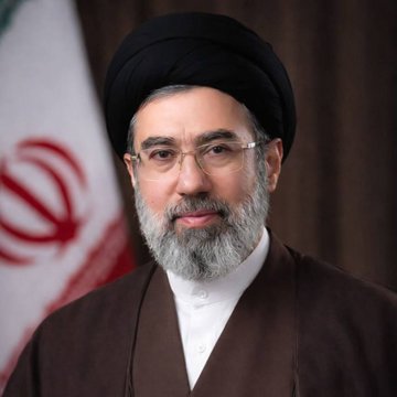 The image features a man wearing traditional Islamic clothing, including a brown robe with a white collar and a black turban. He has a beard and glasses, giving him an intellectual appearance. The background of the photo includes a flag from Iran, which is red and white in color. There are also some text elements present in the image that read "Iran closes the Strait of Hormuz Pray for $BTC to hold".
