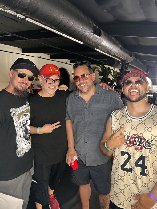Four men pose together indoors in a casual setting with exposed ductwork; one wears a green-patterned hat, another a red cap, a third a grey shirt and sunglasses, and the fourth a beige Gucci jersey with 'KC 24' and a red-patterned headband.