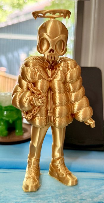 A glossy gold 3D-printed skeleton figure wearing an oversized puffer jacket, chain necklace, and boots stands on a blue surface, matching the "Gorgez" collectible design referenced in the post. The print showcases the bold, streetwear-inspired aesthetic of the design, highlighting the printing quality being promoted from @professorznft at @AtoZNetwork26.