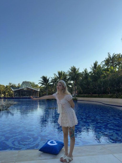 A blonde woman in a light pink dress stands by a large resort pool with palm trees, holding her hand out gracefully with a blue cushion beside her.