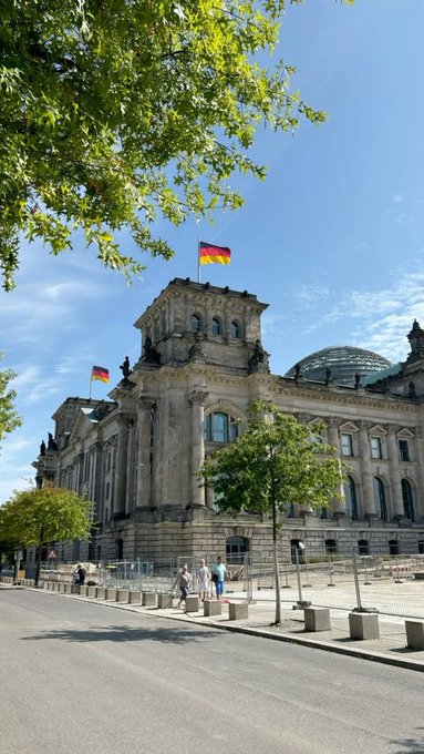 The Reichstag building in Berlin stands under a clear blue sky with German flags flying from the rooftop, partially framed by green tree foliage.