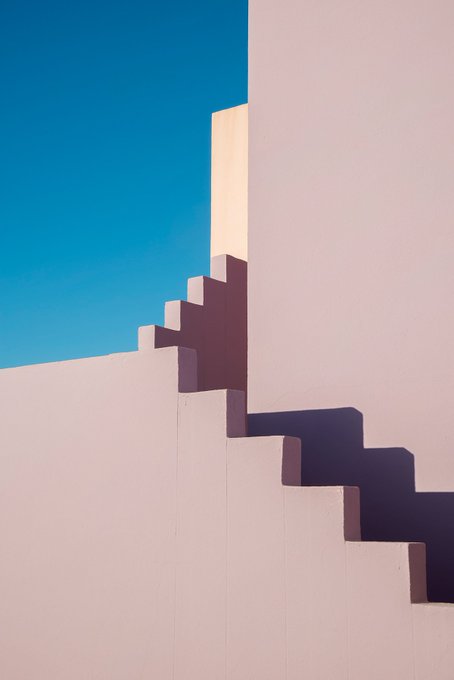 A minimalist architectural photograph showing a pale pink stepped wall casting sharp, dark shadows against a clear blue sky.