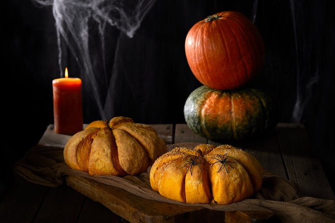 A Halloween-themed still life showing baked pumpkin-shaped pastries and small pumpkins on a wooden board, accompanied by a lit red candle and drifting smoke.