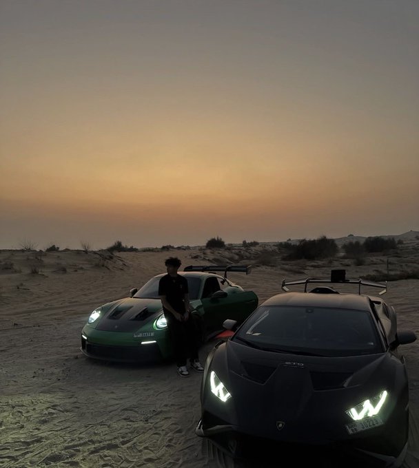 A young man standing between a green Porsche 911 GT3 and a black Lamborghini Huracán STO on sand dunes at sunset.