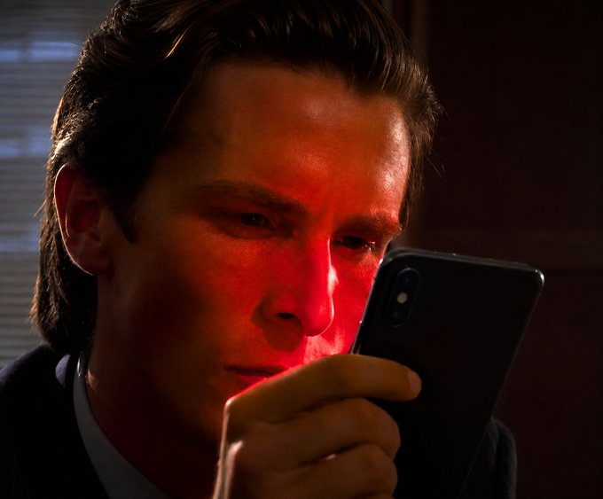 A man in a suit intently stares at a smartphone with a dramatic red glow illuminating his face. The image evokes the "Patrick Bateman checking stocks" meme, humorously paired with a crypto-related post about a Hyperliquid chart that resembles an exciting announcement.