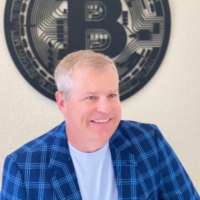 A middle-aged man with short gray hair smiles in front of a wall mounted with a large Bitcoin logo, fitting the podcast episode's focus on Bitcoin mining topics.