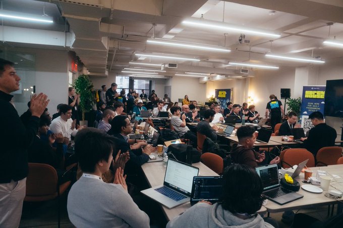 A crowded indoor event space filled with attendees working on laptops at long tables, with yellow 'HACK' branding visible on banners, suggesting a hackathon or tech conference.