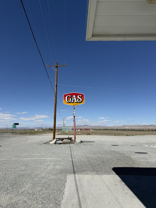 A vintage "GAS" sign mounted on a red pole stands in a barren desert parking lot under a clear blue sky, evoking a sense of remote isolation. The empty landscape and lone utility pole reinforce the "Mobbing" theme of solitary stillness in an abandoned setting.