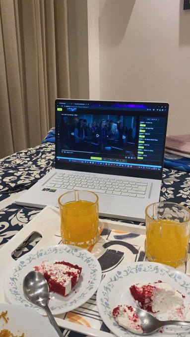 A laptop on a bed plays a video, with two glasses of orange juice and two small plates of red velvet cake in the foreground.