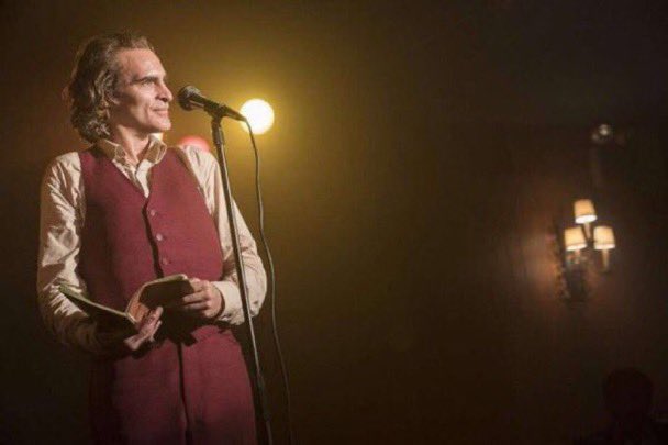 A man in a vest stands at a microphone, holding a book or notes, as if performing a spoken word piece or reading on a warmly lit stage. The moody, contemplative atmosphere of the image echoes the reflective sentiment in the post about Sui having reached its bottom.
