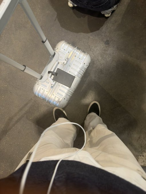 A first-person view looking down at a silver Rimowa suitcase on the floor in front of the person's feet, with a luggage handle extended.