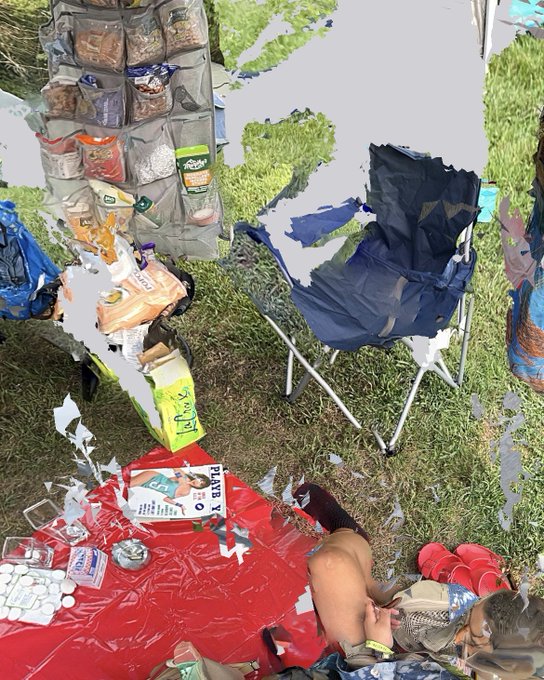 The image shows a person sitting on a red blanket, surrounded by various items such as bags of chips, food, and drinks. The scene appears to be set up for an outdoor gathering or picnic, with chairs and other objects scattered around the area. The photo is taken from a low angle, giving prominence to the person sitting in the center of the image while also capturing the surrounding elements.
