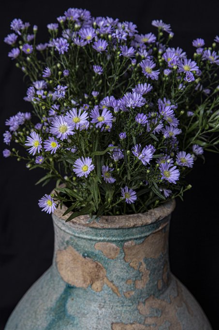 A bouquet of purple aster flowers with bright yellow centers sits in a weathered turquoise ceramic vase with patches of exposed clay, set against a dark background. The image appears to be an art piece being sold in the secondary market, with the seller thanking a buyer and congratulating others in the art community.