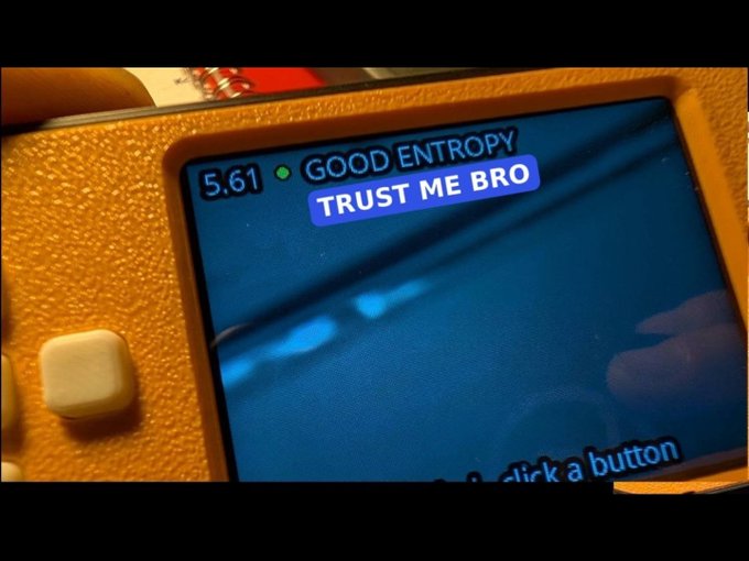 A close-up of a yellow handheld gaming console screen displaying '5.61 GOOD ENTROPY' and 'TRUST ME BRO'.
