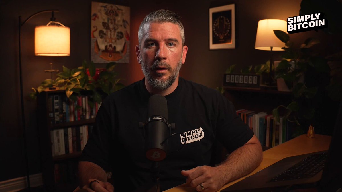 The image shows a man sitting at a desk with a microphone, wearing a black shirt that says "Stroke Bitcoin" on it. The background features two lamps and several bookshelves filled with various books. There are also potted plants in the room, one near the left side of the scene and another towards the right side.