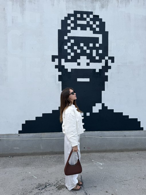 A woman in a white outfit and sunglasses stands in front of a large pixel-art mural of a face, carrying a brown bag.