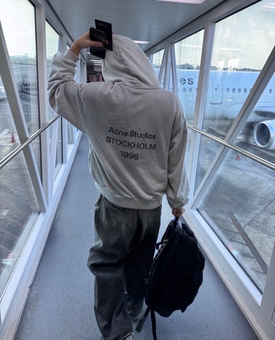 Photo of a person from behind walking through an airport jet bridge, wearing a gray hoodie reading 'Acne Studios STOCKHOLM 1996' and carrying a black backpack, with an airplane visible outside.