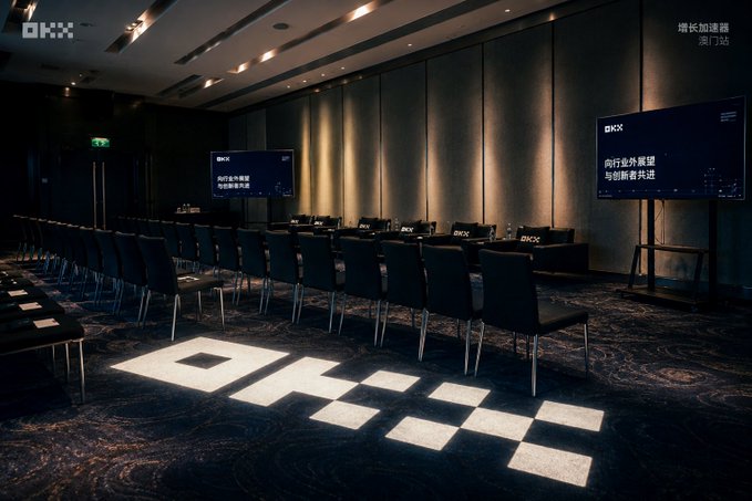 A dimly lit conference room with rows of black chairs facing two screens displaying OKX logos and Chinese text, with the OKX logo projected onto the carpeted floor.