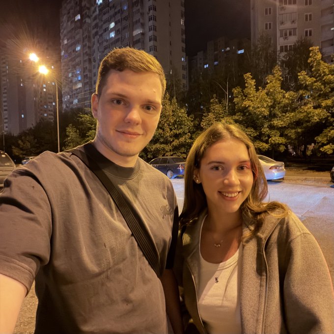 A nighttime selfie of two smiling young people in an urban setting with illuminated apartment buildings and parked cars in the background. The image accompanies a post about meeting online connections in person, capturing a real-life meetup in Kyiv between crypto community members.