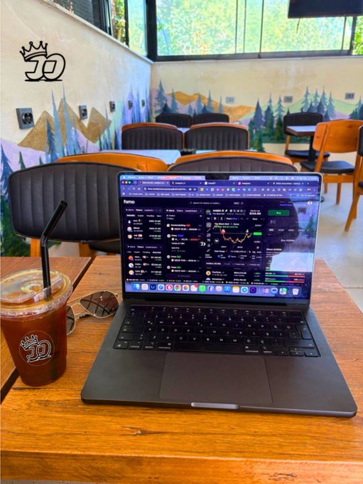 A laptop on a wooden table in a coffee shop displays a trading interface with market data, accompanied by an iced coffee in a plastic cup and sunglasses.
