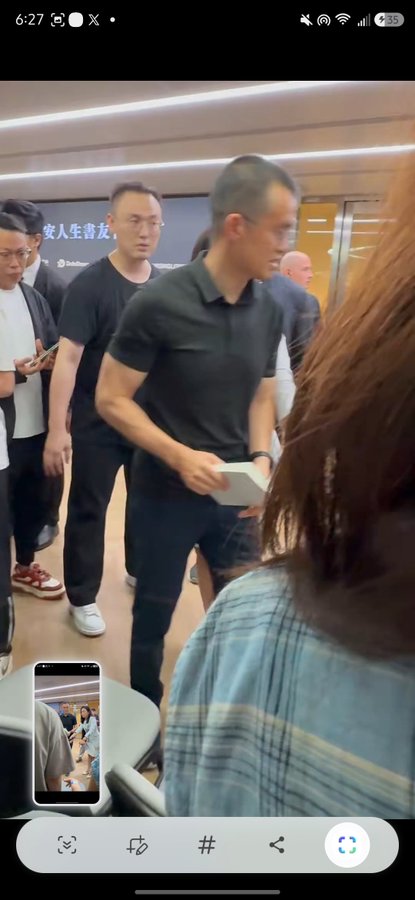 A crowded indoor event with several men in dark clothing, one holding a white box, while a woman in a plaid shirt stands in the foreground; Chinese text "安人生喜友" is visible on the wall, and a phone screen in the corner shows another angle of the gathering. The image captures a bustling scene that the post seems to be inquiring about, possibly seeking context or identification of the event.
