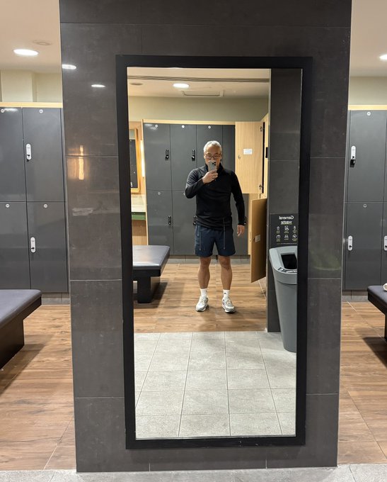 A man takes a mirror selfie in a locker room, wearing a black long-sleeve shirt, blue shorts, and white sneakers.