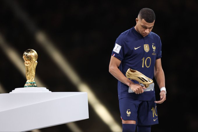 Kylian Mbappé wearing France's #10 jersey walks past the FIFA World Cup trophy, holding his golden boot award and looking down dejectedly.