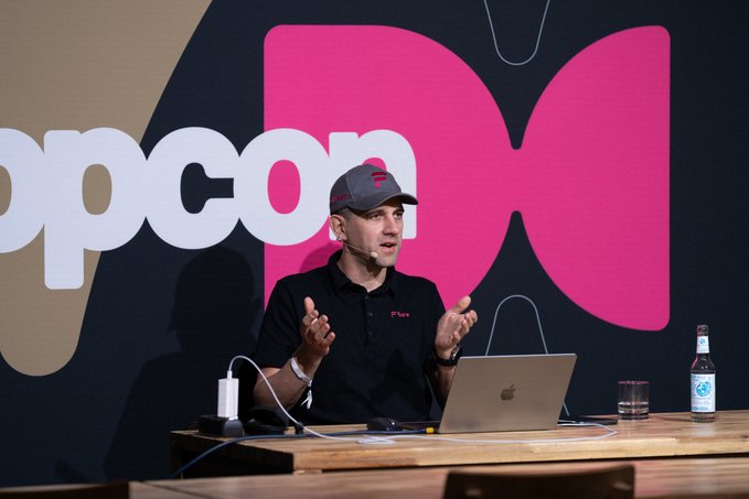 Man in a black polo shirt and cap speaking at a conference table with a laptop, in front of a large 'opcon' logo backdrop.