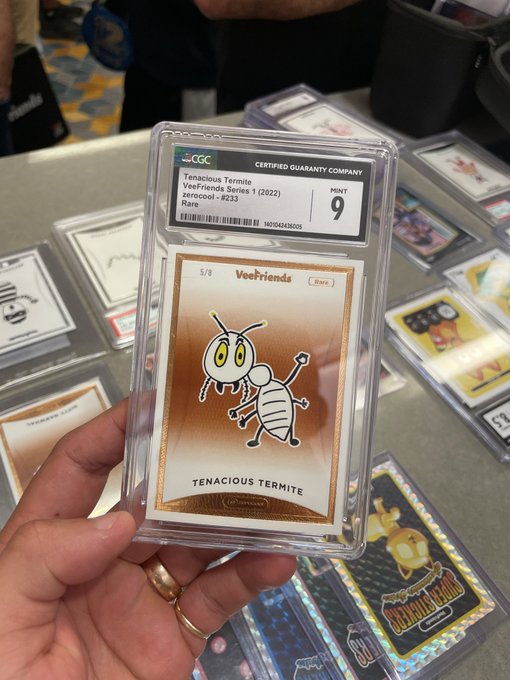 A hand holds a CGC-graded Mint 9 'Tenacious Termite' VeeFriends Series 1 trading card, featuring a cartoon termite and numbered 5/8, at what appears to be a collectibles event with other graded cards displayed nearby.