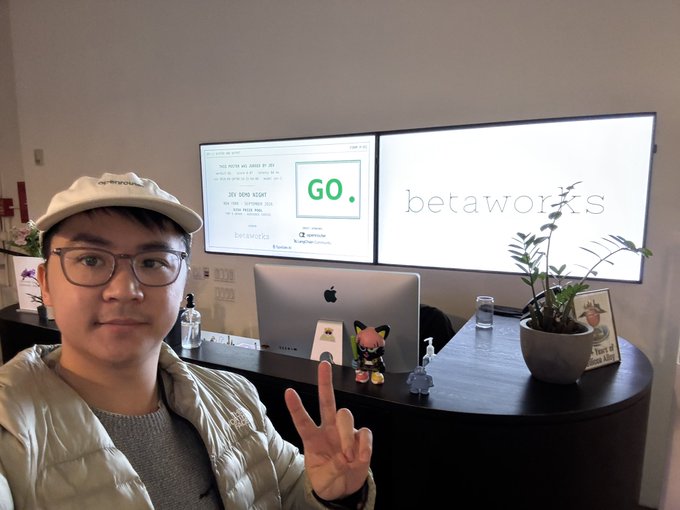 A selfie of a young man wearing a beige cap, glasses, and a puffy jacket, making a peace sign at an office desk with dual monitors displaying 'GO.' and 'betaworks'.