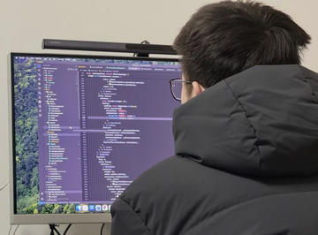 A person in a dark puffer jacket sits facing a large monitor displaying lines of code, with a lightbar attached to the top of the screen.