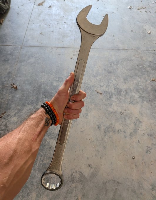 A hand adorned with beaded bracelets grips a massive combination wrench stamped "60mm," its imposing size a literal embodiment of the metaphor that "sovereignty requires maintenance"—just as the oversized tool demands strength to wield, self-rule demands continual effort and upkeep.
