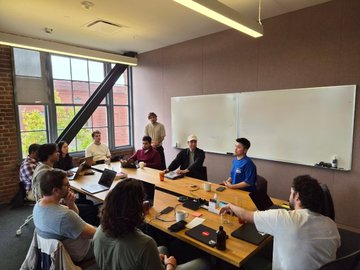 Two members of Holdstation are seated at a conference table during a meeting in a modern office space with a whiteboard, where they appear to be presenting or collaborating with other participants on a laptop.