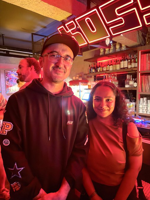 A man wearing a black hoodie and cap stands next to a woman in an olive top inside a bar with a neon 'LOSER' sign overhead.