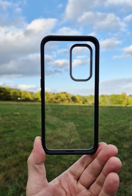 A hand holding up a transparent phone case with a black border against a backdrop of a grassy field, trees, and cloudy sky.