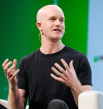 A man in a black t-shirt speaks on a stage with a green screen backdrop, gesturing expressively with both hands.