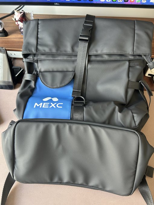 A gray backpack featuring a blue front pocket with the white "MEXC" logo rests on a desk beneath a Dell monitor.