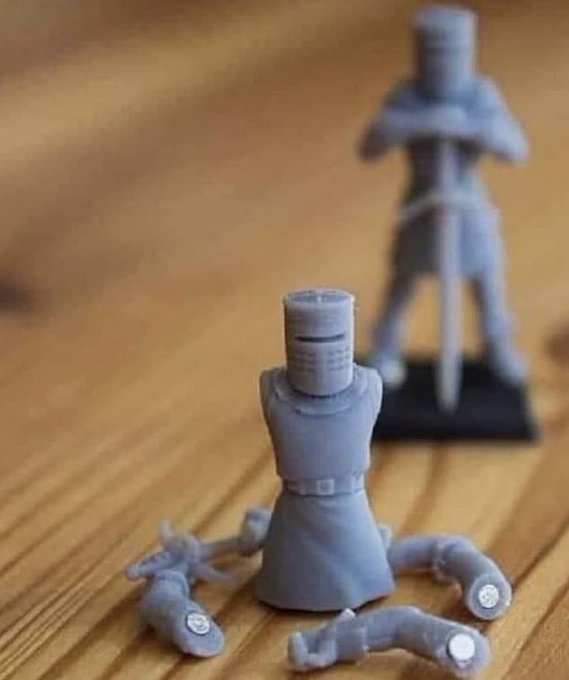 3D-printed grey plastic knight figurines on a wooden surface, with one sitting in the foreground surrounded by scattered armor pieces while another stands in the background.