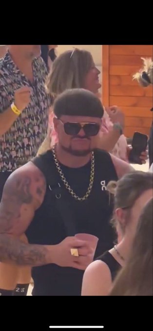 A photo of a man with a bowl haircut, sunglasses, a goatee, and tattoos, wearing a thick gold chain and black vest, holding a pink cup in a crowded, social setting.