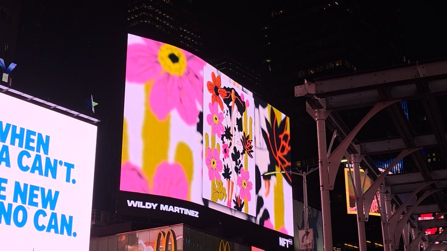 A nighttime view of illuminated billboards in Times Square, including one with bright pink flowers and text crediting 'WILDY MARTINEZ', alongside a sign with blue text reading 'WHEN / CAN'T. / NEW / CAN.'