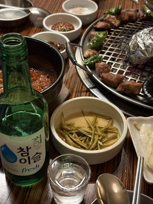 A green bottle labeled "참이슬 fresh" (Korean soju) and a small glass of clear liquor sit beside a Korean BBQ spread of grilled meat on a wire mesh, side dishes, and pickled greens, celebrating a casual drink to mark Ethereum's rise.