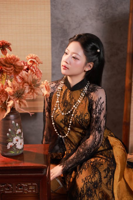 A portrait of a young woman with long black hair adorned with a pearl hairpin, wearing a gold and black lace cheongsam with pearl necklaces, sitting next to a vase of orange flowers on a wooden table.
