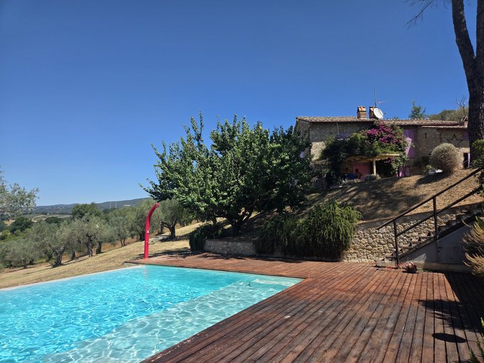 A scenic view of a stone house with a wooden deck and swimming pool in the foreground, set in a rural landscape with olive trees and a clear blue sky.