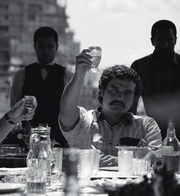 A black and white photo shows a mustachioed man raising a small glass in a toast while seated at a table with empty dishes and glasses, flanked by two other men in the background.