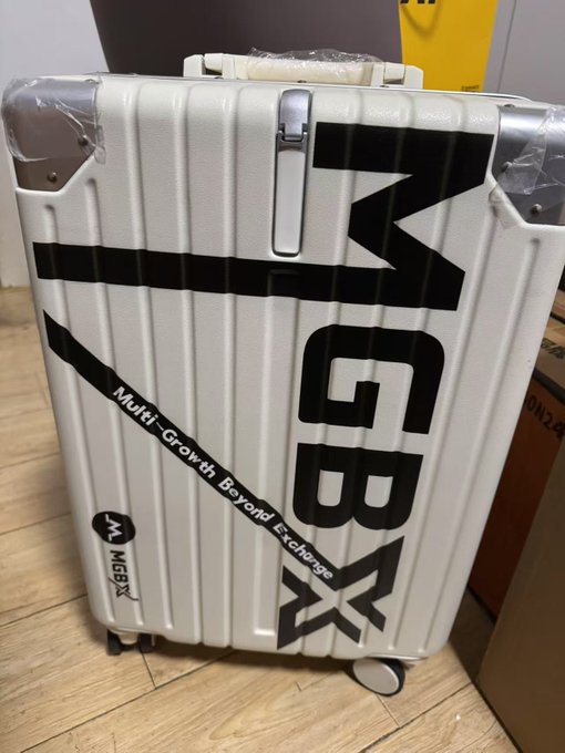 The image features a white suitcase with black text that reads "MGBX" and "MGBX_ZH @MGBX_ZH @Heidi_MGBX". The suitcase appears to be new, as indicated by its pristine condition. It is placed on the floor next to some boxes, suggesting it might have been recently purchased or is waiting for transportation.