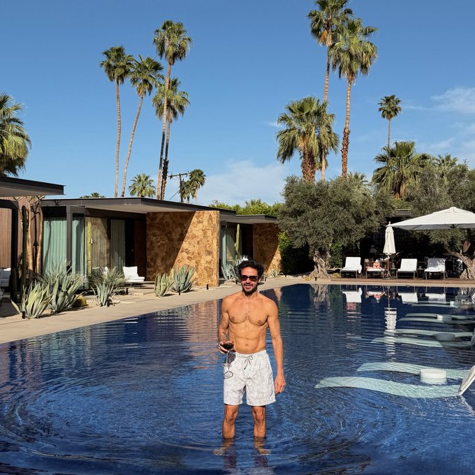 The image features a man standing in a swimming pool with his arms raised above his head. He appears to be enjoying himself, possibly taking a break from swimming or posing for a photo. The surrounding area includes palm trees and other buildings, creating an outdoor setting. There is also text present that reads "Hey, what's up! I'M A SON OF THE BILLION!". This message seems to be related to the man in the pool, possibly indicating his status as a Bitcoiner or someone who has achieved significant wealth through cryptocurrency investments.
