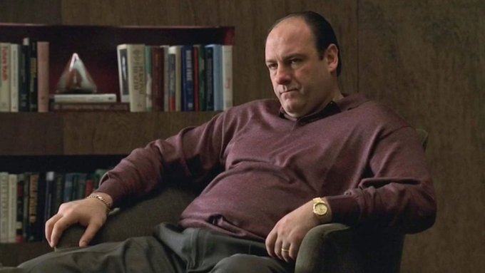 A balding, heavyset man in a maroon long-sleeve shirt sits slouched in a leather chair in front of a bookshelf, his expression deadpan and slightly uncomfortable as if admitting defeat—mirroring the post's dialogue about trading high-leverage perpetuals while avoiding "complicated" options despite repeated liquidations.