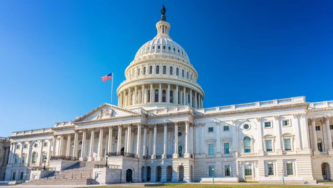 The image shows the United States Capitol Building, featuring its iconic white dome and neoclassical architecture, with an American flag flying against a clear blue sky. This visual relates to the breaking news about the delayed CLARITY Act vote, as the Capitol is the symbolic center of U.S. legislative activity where such bills would be considered by the Senate upon their return from recess.